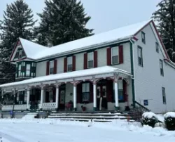 A two-story white house with a covered porch, red shutters, and evergreen trees, all covered in snow.