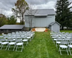 A wedding ceremony setup with rows of white chairs on a lawn in front of a rustic barn.