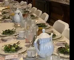 dining table set with white plates, silverware, and a large white coffee or teapot.
